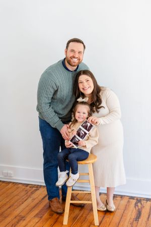 A family poses together in front of a white wall while holding an ultrasound photo on a wooden stool.