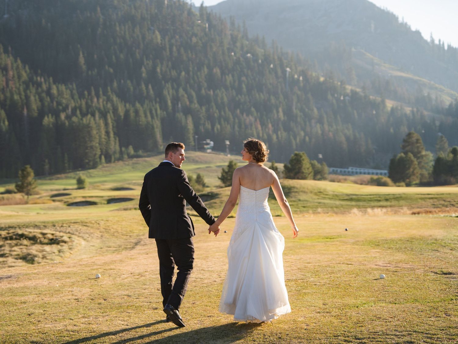 Couple in wedding attire walking hand in hand along a scenic beach with mountains and water in the background.