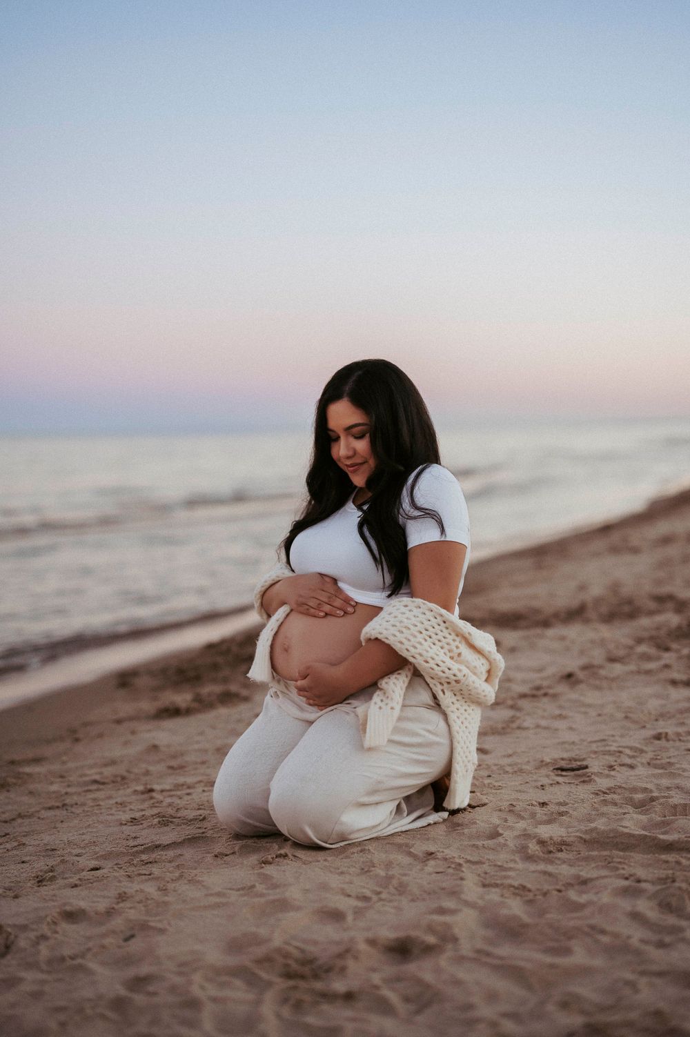 Expectant mother in white outfit kneeling on beach at sunset.