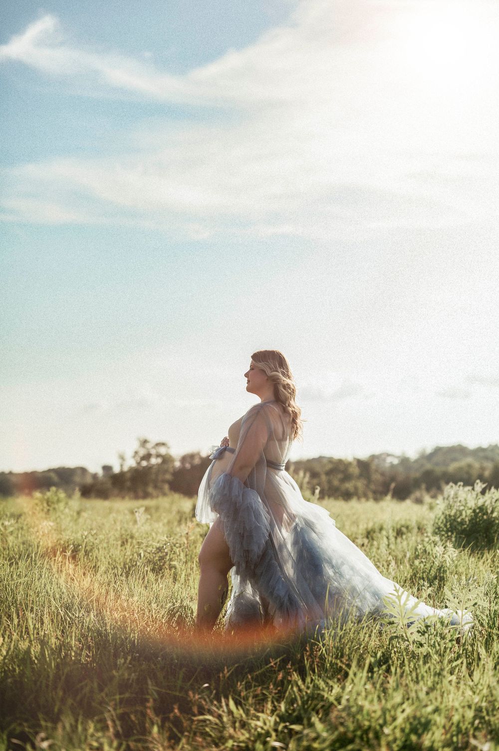 Ethereal maternity portrait of woman in flowing dress standing in sunlit meadow.