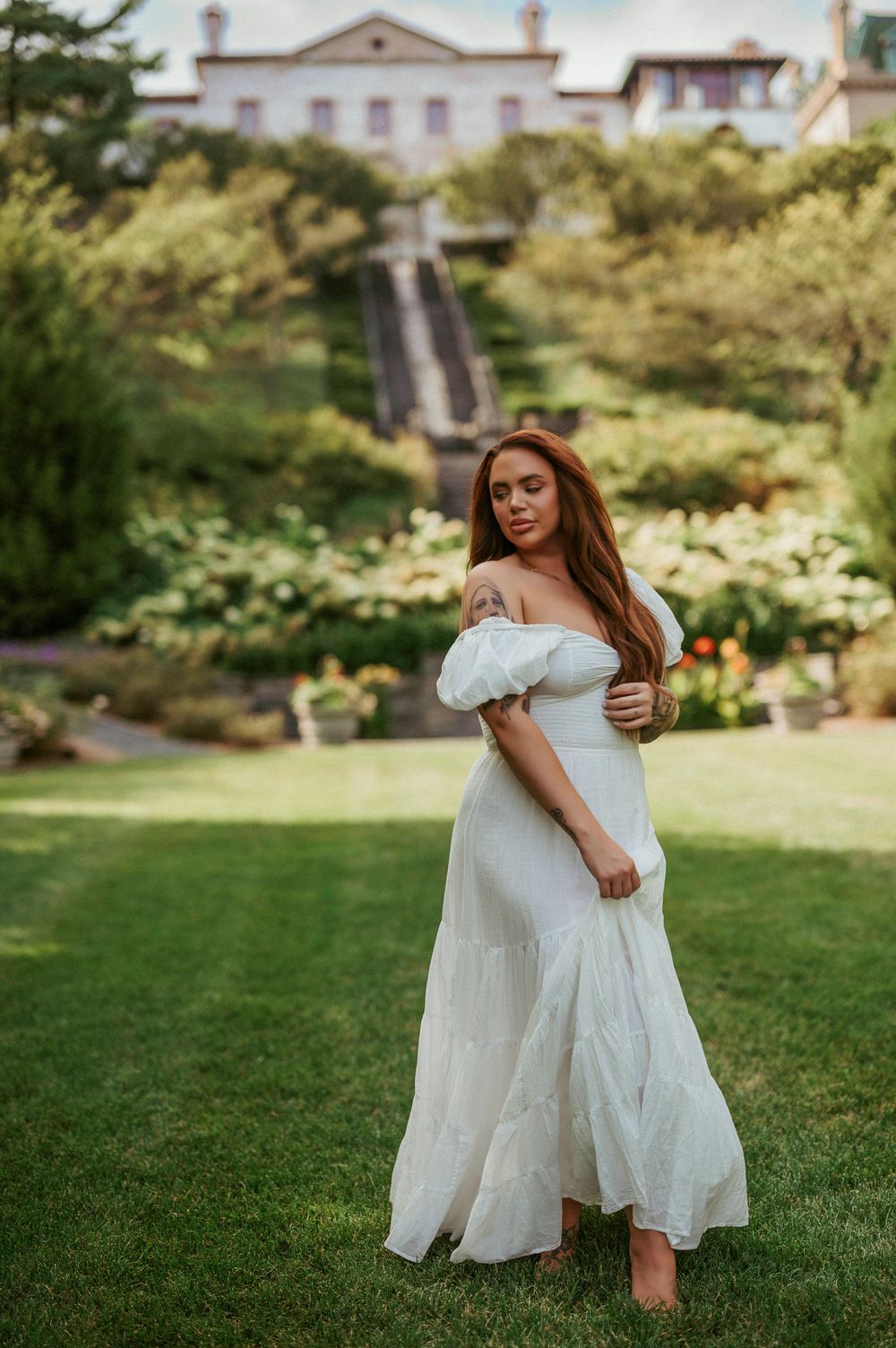 Woman in flowing white dress poses in garden with mansion and staircase in background.