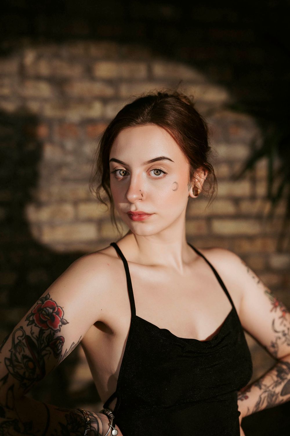 Moody portrait of tattooed woman in black tank top against brick wall.