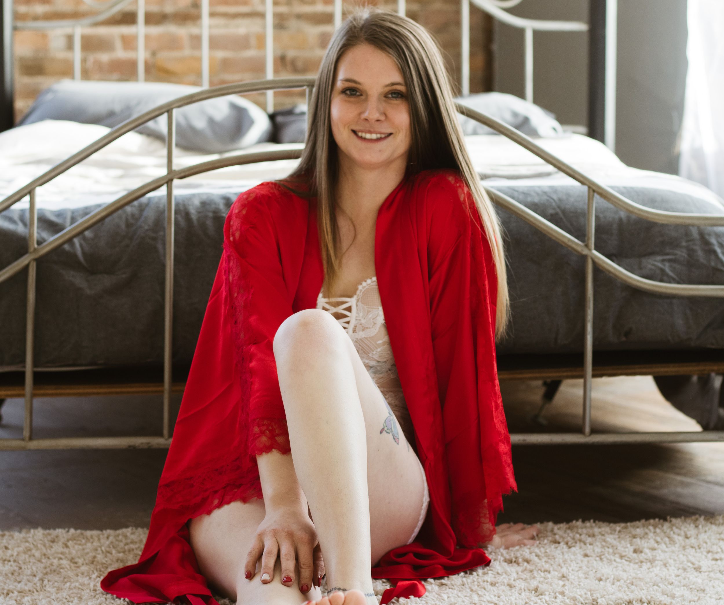 Person in long red robe sitting casually on carpet next to bed wearing light colored clothing.