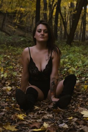 A moody fall scene in a forest shows a figure sitting among scattered autumn leaves wearing black lace and boots.