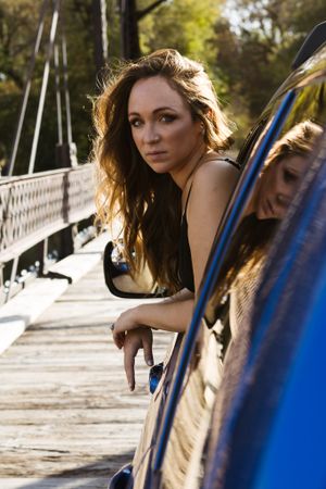 Woman with windswept hair leans against blue car on wooden bridge in warm sunlight.