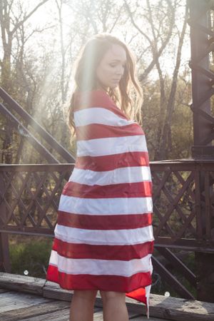 A person wrapped in a red and white striped American flag stands on a wooden bridge at sunset.