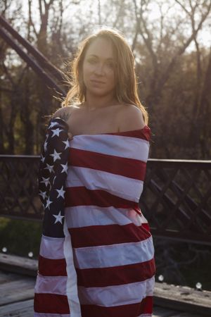 A person draped in an American flag poses against backlighting on a wooden bridge.