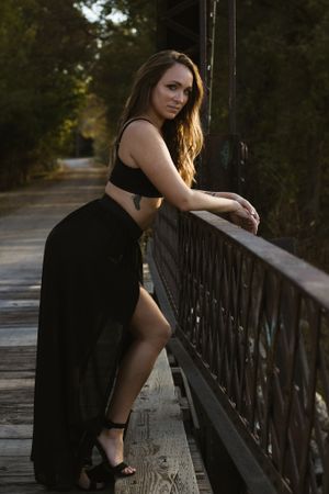 A person in a black dress poses against a rustic bridge railing at dusk.