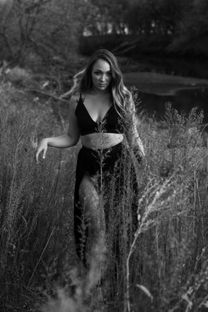 A person in black clothing stands among tall grasses in a moody black and white portrait.