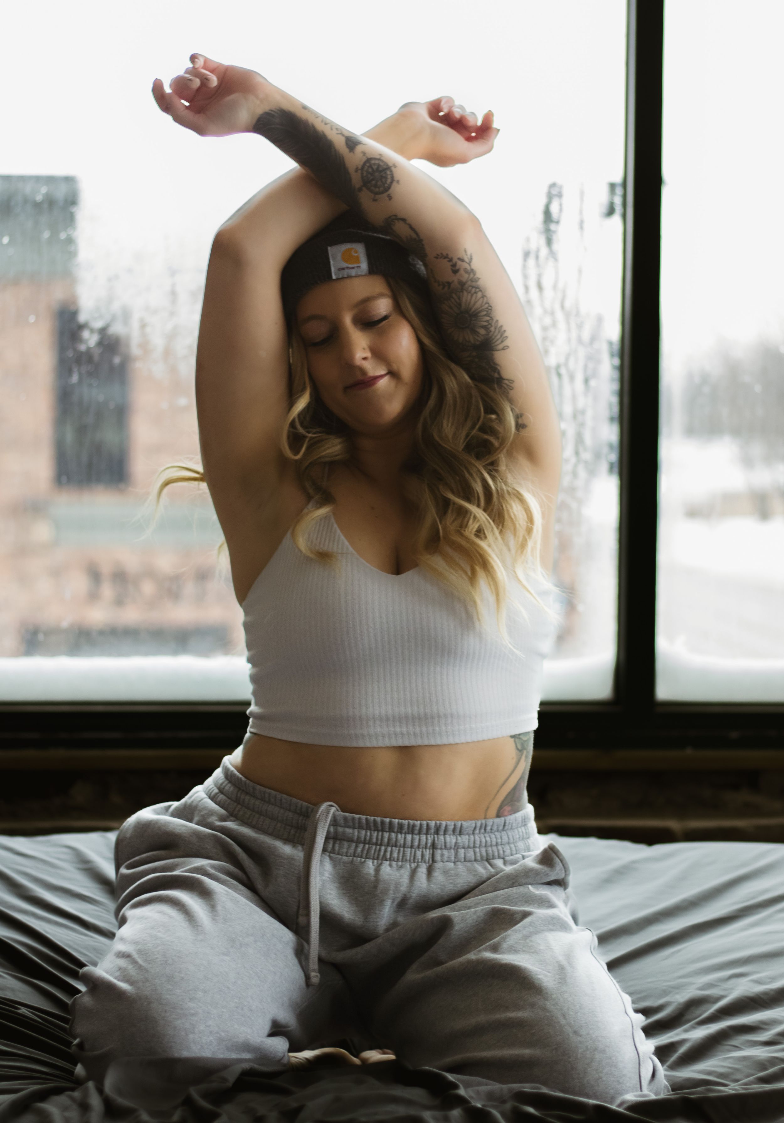 A relaxed figure in a white crop top and grey sweatpants stretches on a bed near a window on an overcast day.