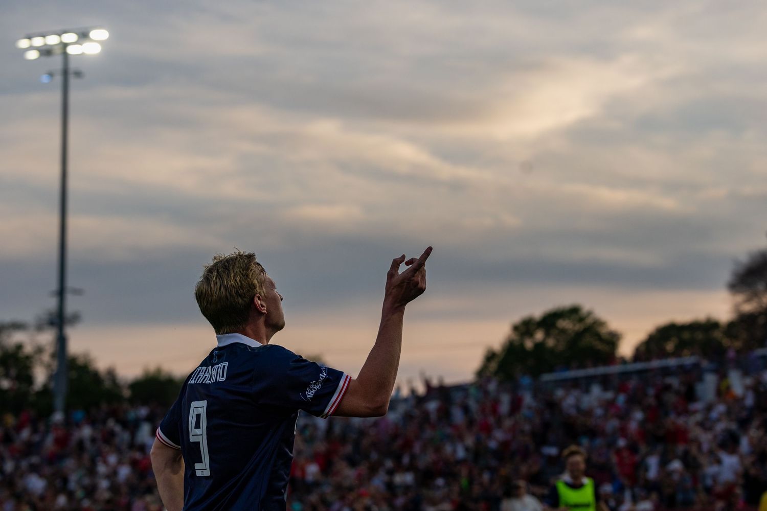 Stadium crowd silhouetted against dramatic cloudy sunset sky.