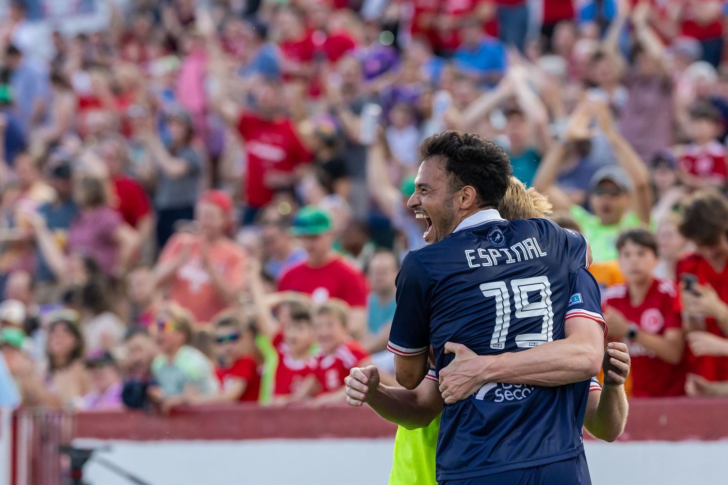 Soccer fans celebrate in stadium stands wearing red and navy jerseys.