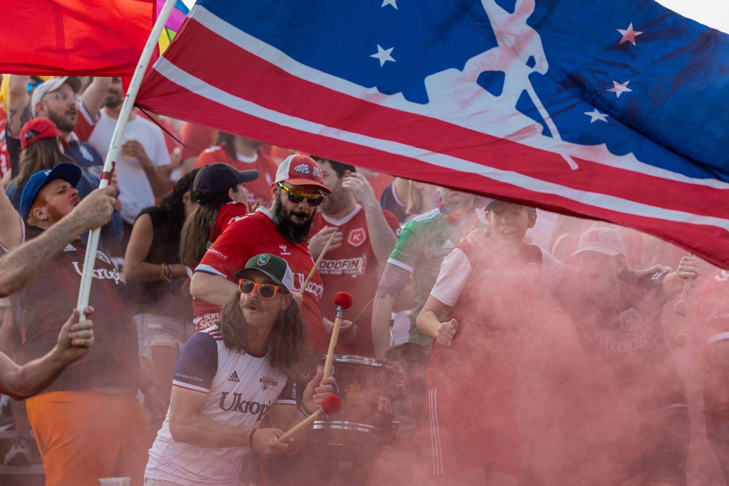 American flag waves above a crowd with red smoke effects at a sports event.