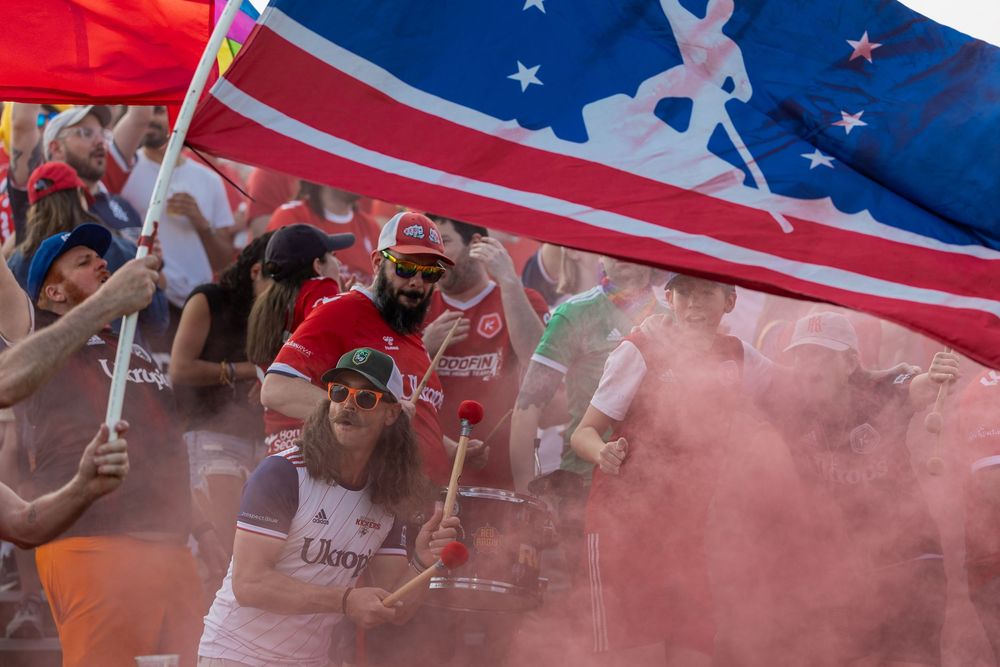 American flag waves above a crowd with red smoke effects at a sports event.