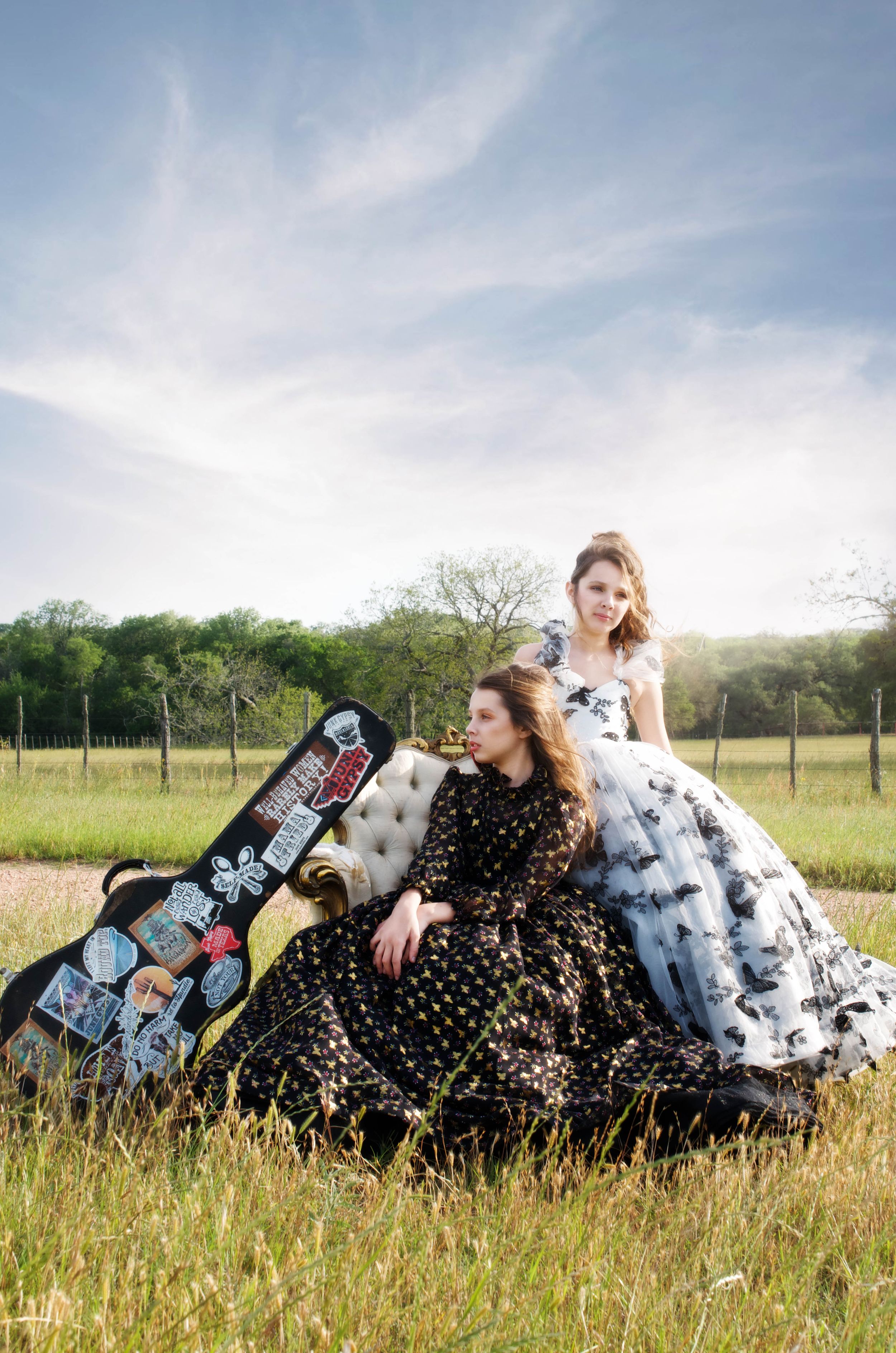 Two sisters in vintage-style couture dresses pose in a grassy field with a guitar case decorated with patches on a sunny day.