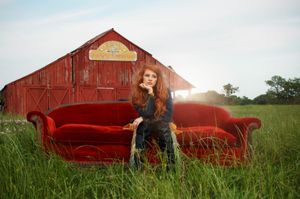 A red headed girl sits on red velvet couch in a grassy field at sunset with a rustic red barn in the background.