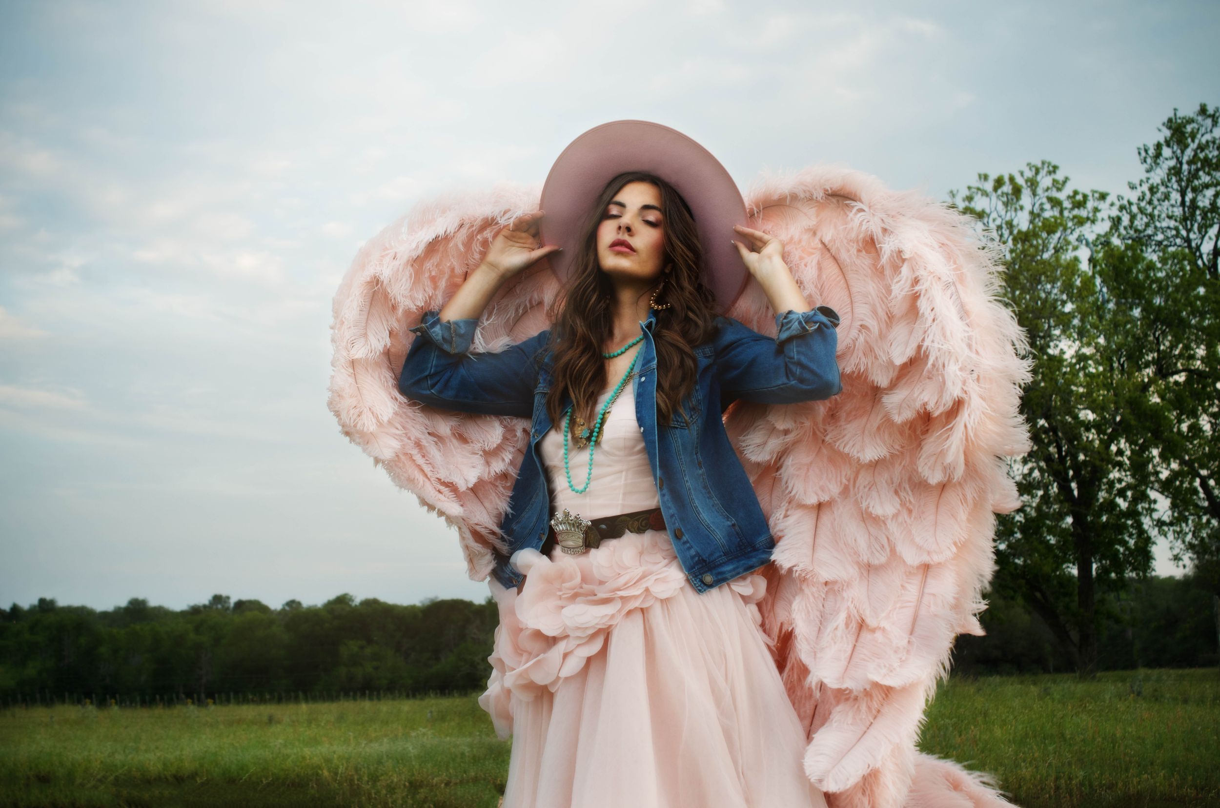 A girl in a pink Vera Wang dress, denim jacket, and couture angel wings twirls gracefully in a meadow in Texas.