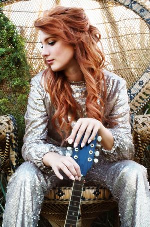 A girl in a sparkly silver jumpsuit sits with a guitar in an outdoor setting in a wicker peacock chair during golden hour.