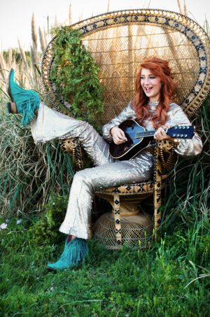 A photograph showing a red-haired girl in a silver sequin jumpsuit and turquoise boots lounging playfully on a wicker peacock chair.