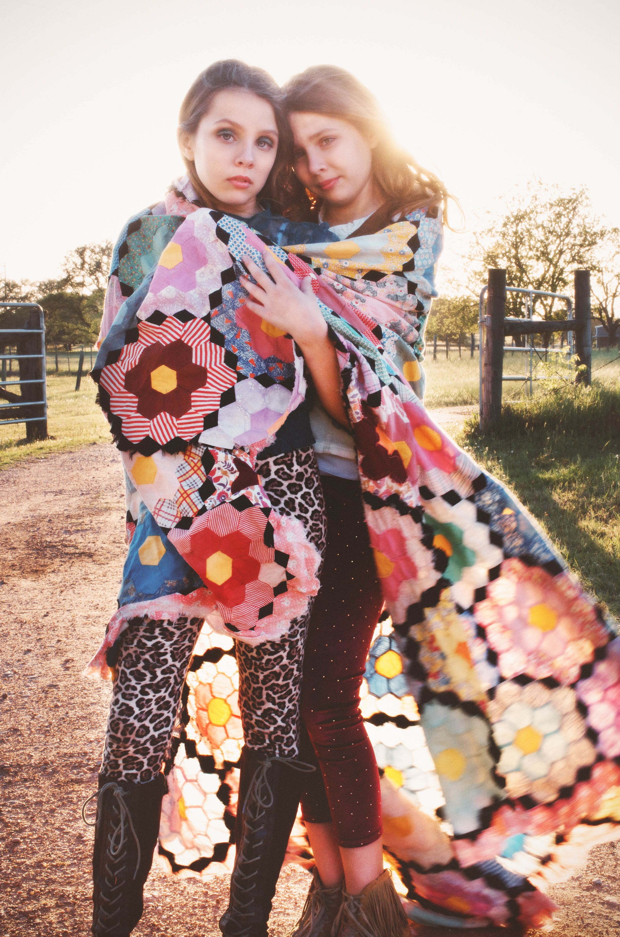 Two sisters stand together wrapped in colorful patchwork quilt with geometric patterns against a sunset field backdrop.
