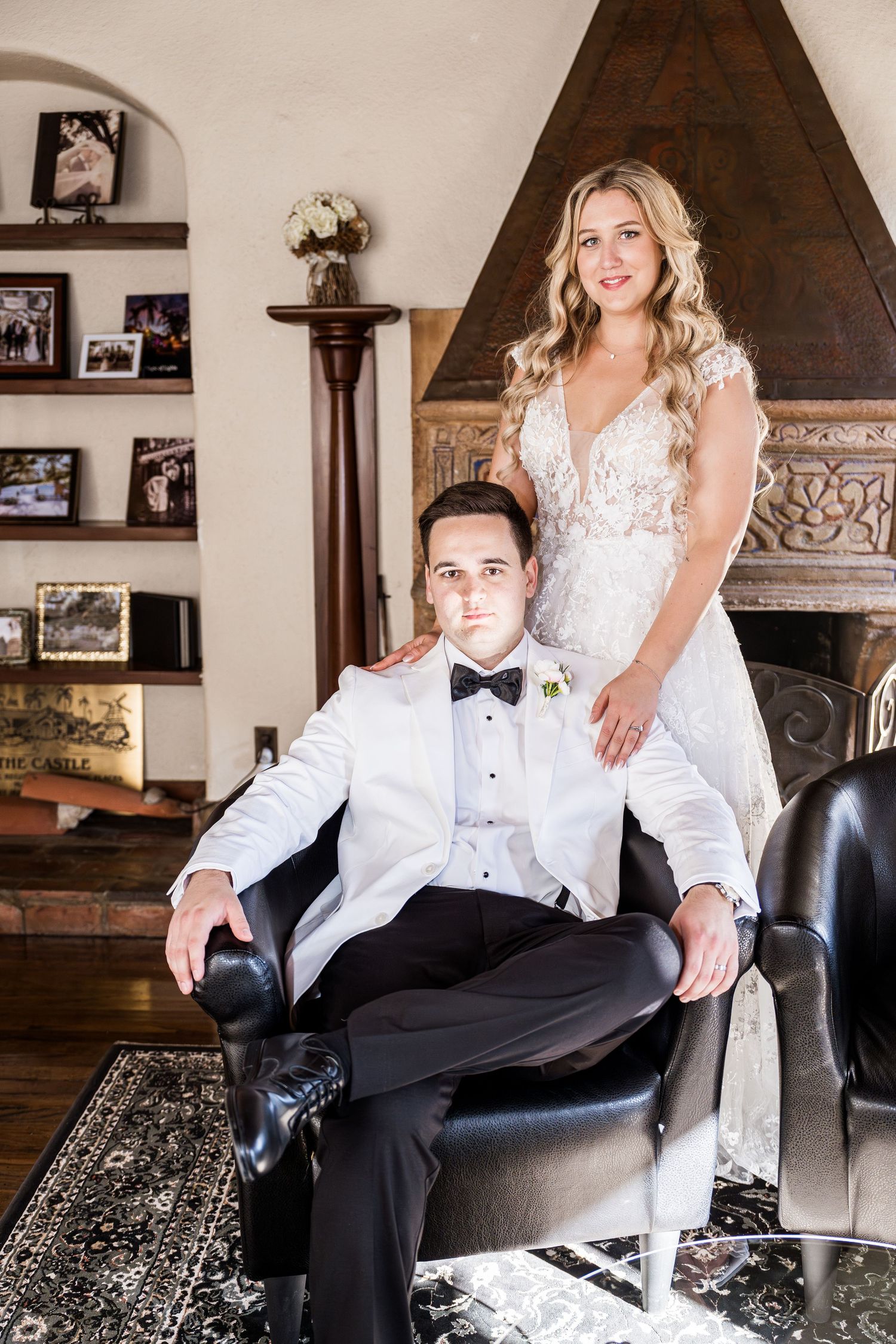 A couple poses together on a black leather chair in a rustic room with wooden beams and framed photos on the wall.