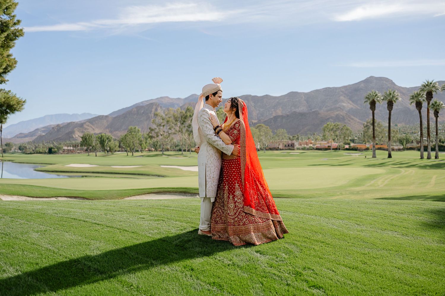 Wedding couple on golf course with mountains, bride wearing traditional red Indian lehenga.