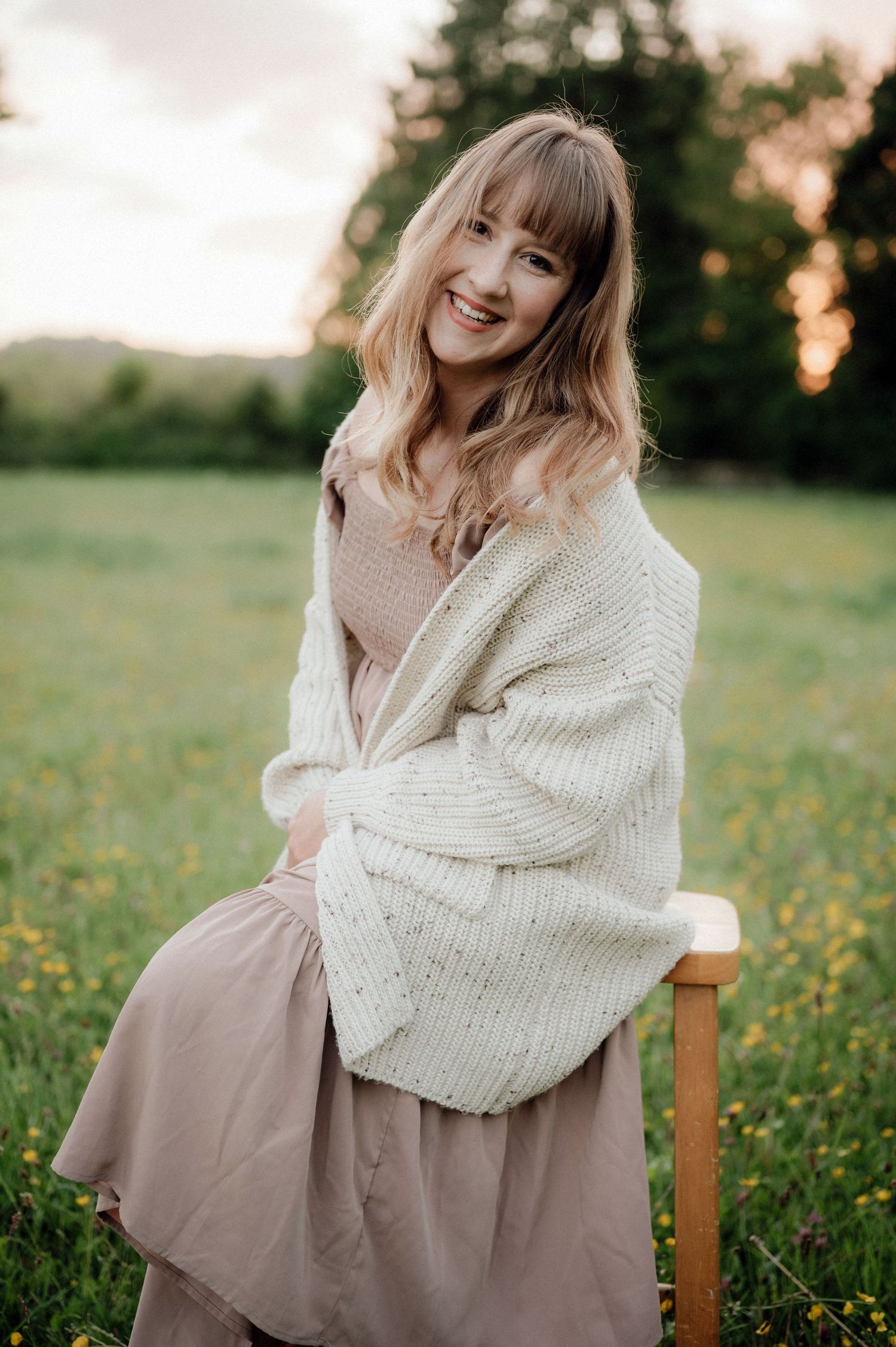 Lisa from Pausing Time Photography in a cosy sweater sits in a wildflower meadow wearing a flowing beige dress in Cheltenham