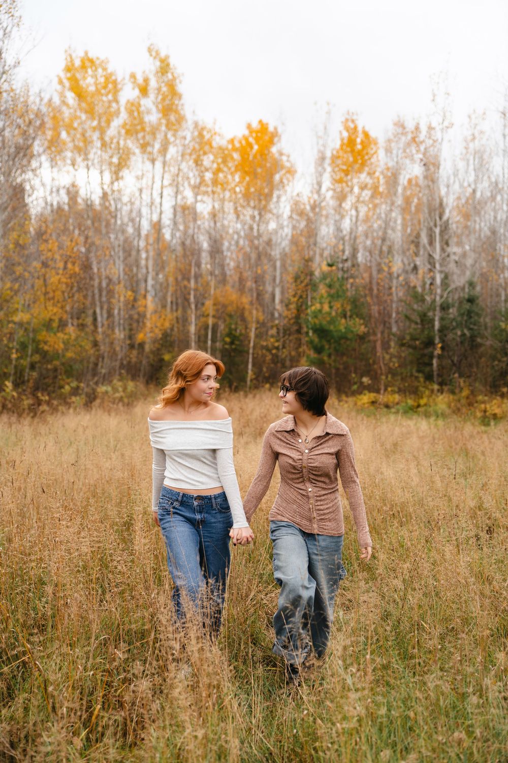 Two people running through a golden autumn field with tall grass and trees in the background during fall season.