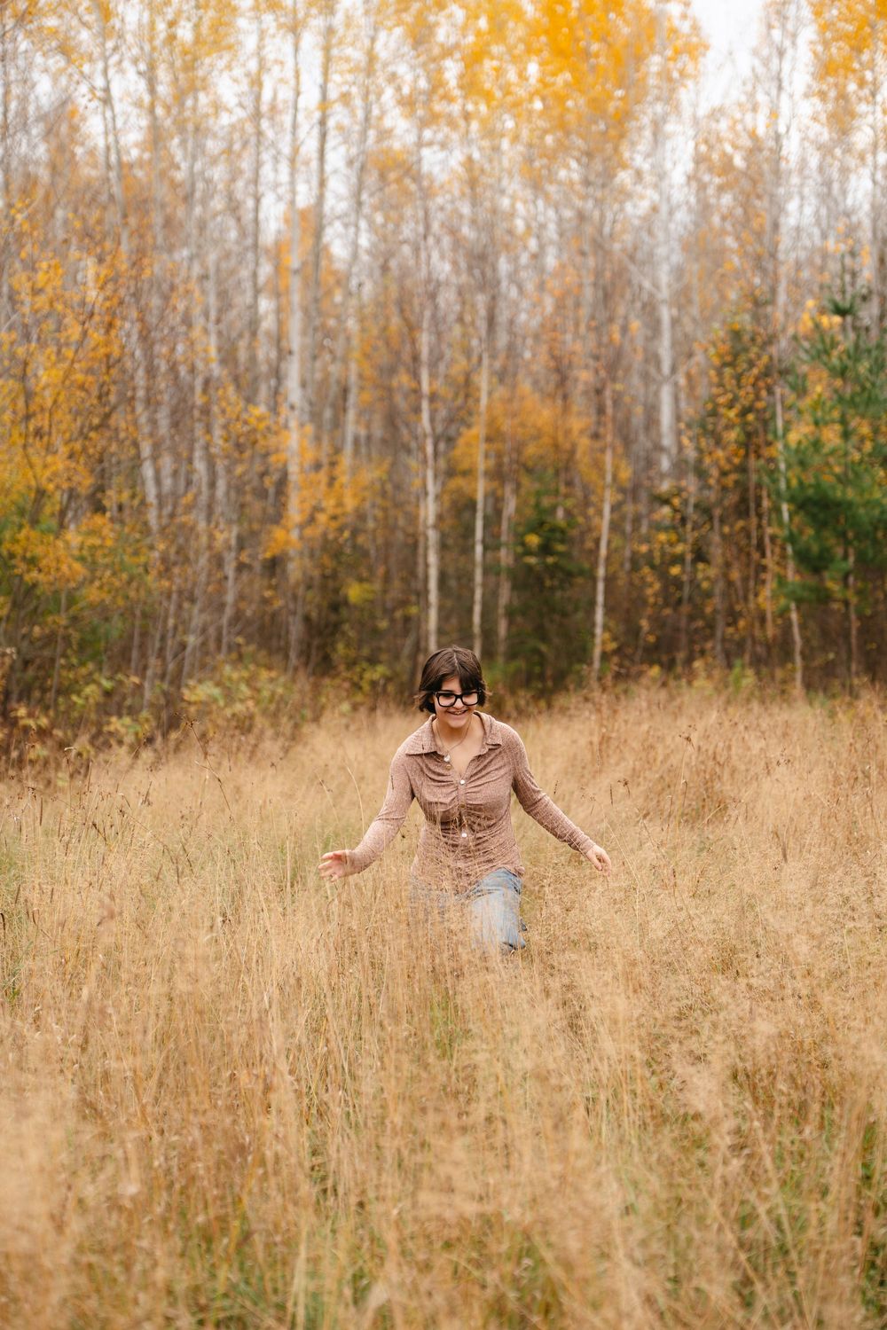 Person frolicking through tall golden grass in an autumn field with birch trees in the background.