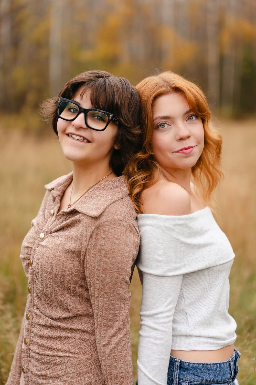 Two friends in casual sweaters pose back-to-back outdoors during autumn with natural fall foliage in background.