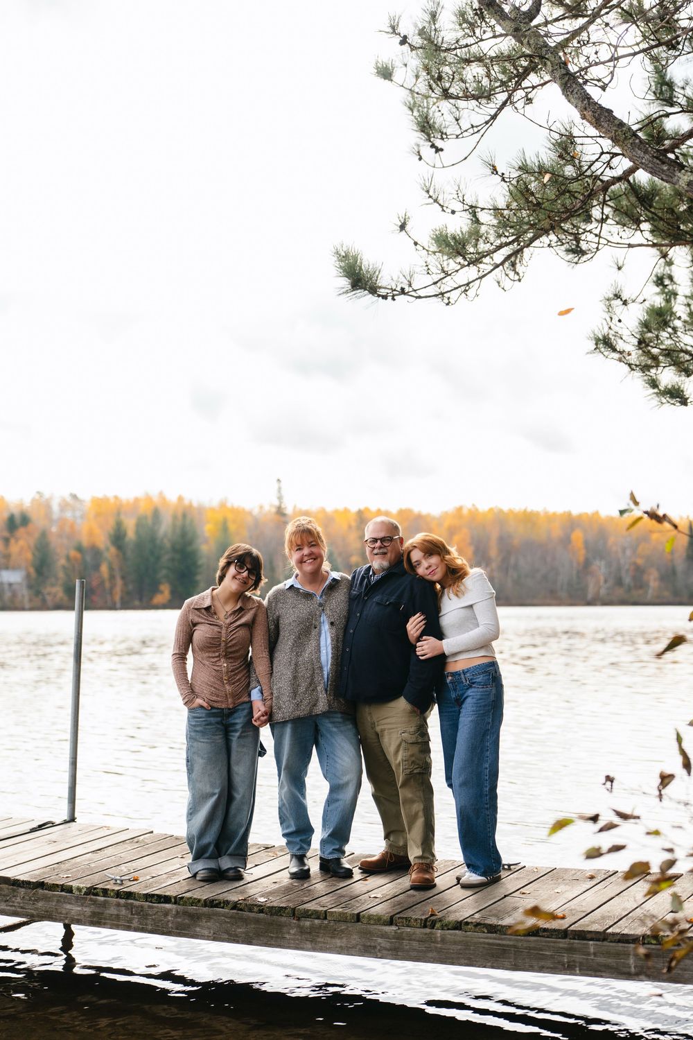 A group of people lean against each other playfully while posing on a wooden dock by a lake during autumn.