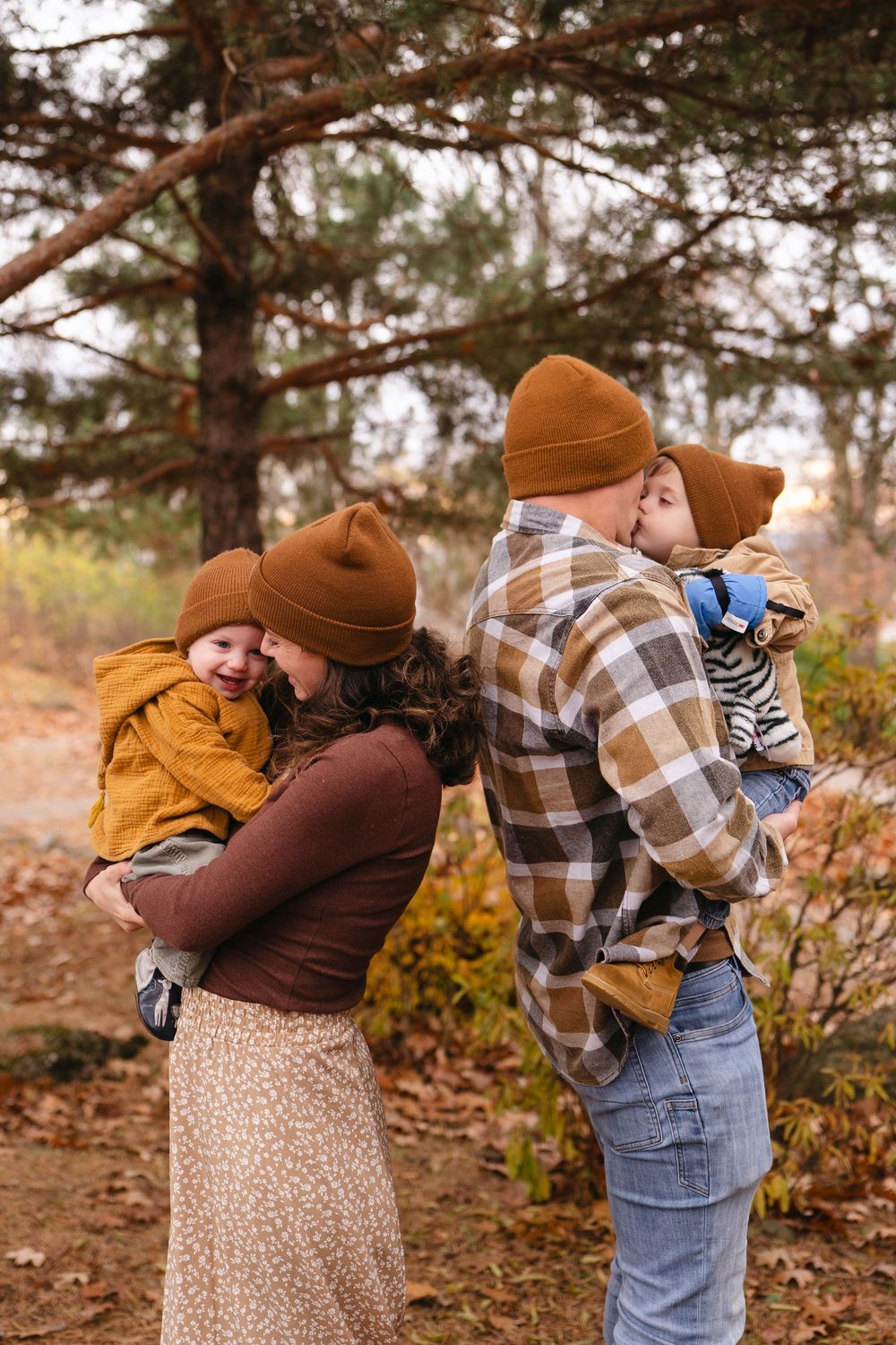 A couple in autumn attire embraces outdoors among fall foliage and pine trees.