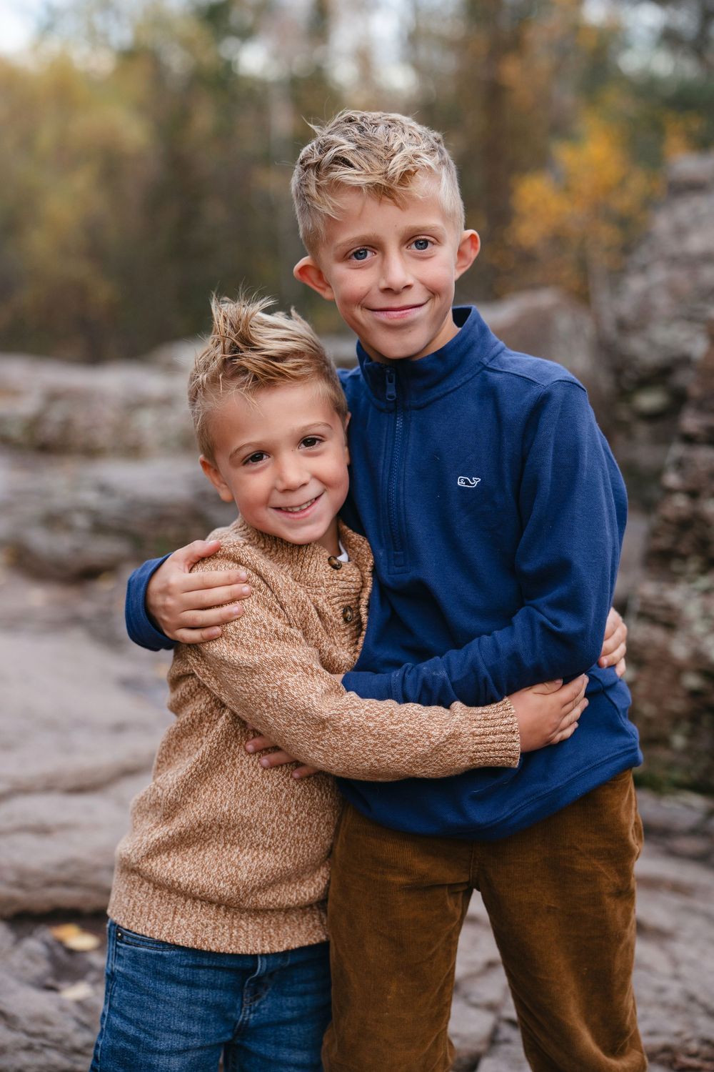 Two children in blue and brown sweaters embrace and smile while posing outdoors in an autumn setting.
