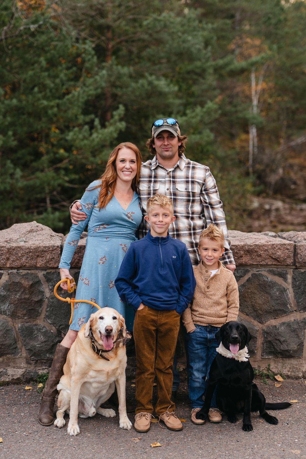 A family stands together with their two dogs on a stone wall path surrounded by autumn trees in a park setting.