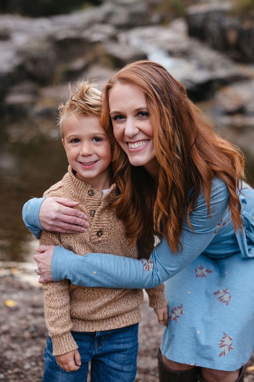 A parent and child share a warm embrace outdoors by a rocky stream, both wearing cozy sweaters on an autumn day.