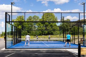 Modern padel tennis court with glass enclosure and players during daytime match.