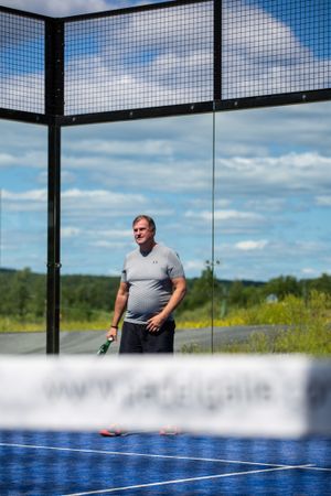 Padel tennis court with glass walls and player watching game against blue sky.