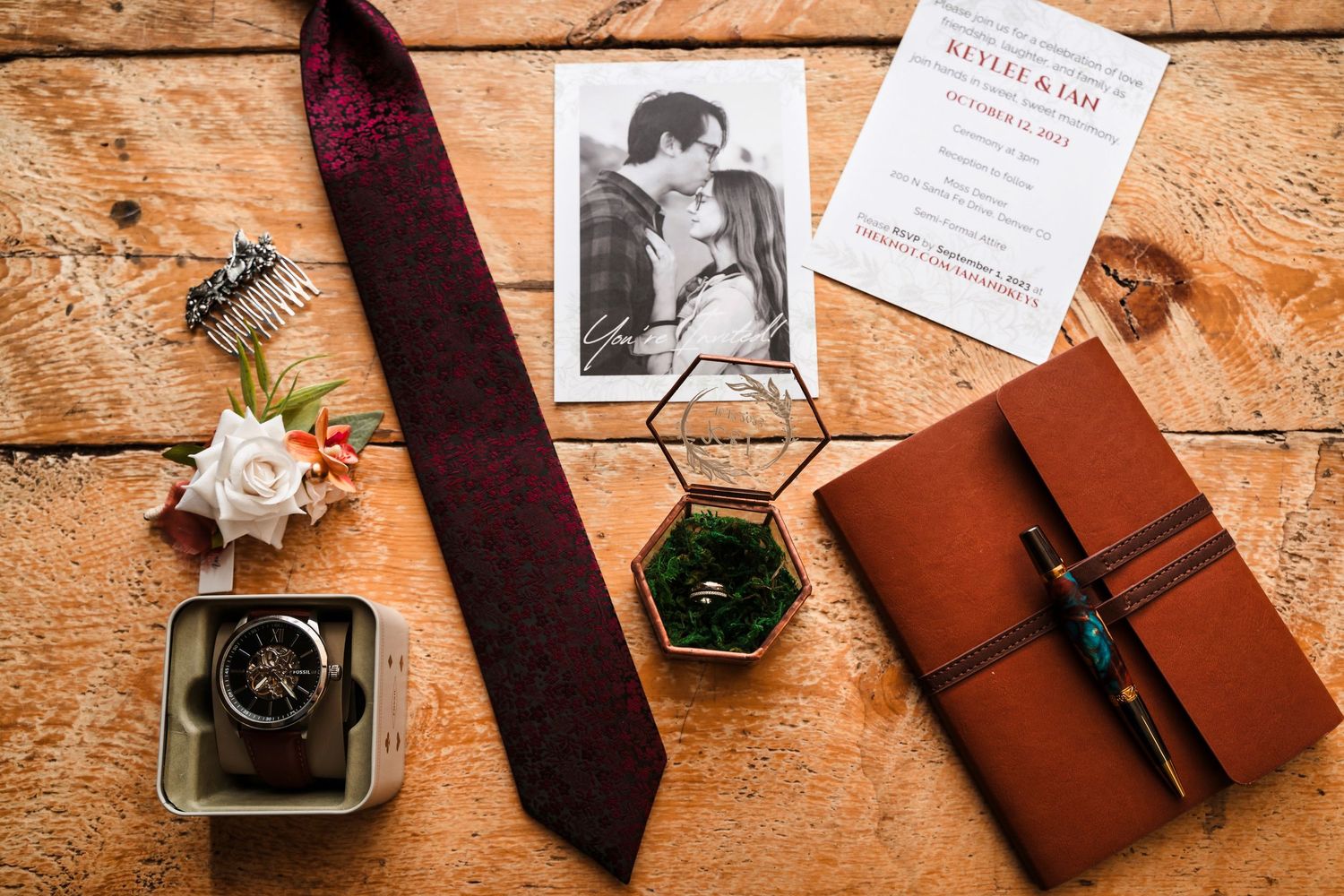 Wedding accessories including a burgundy tie, vintage photograph, leather journal, and green ring box on wooden surface.