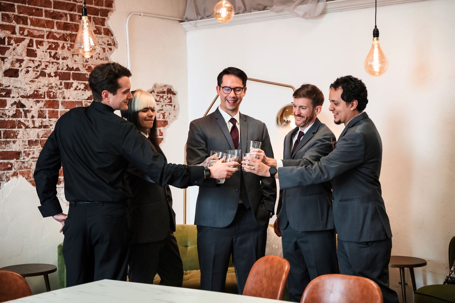 A group of men in suits clink champagne glasses together in celebration at an indoor venue with exposed brick walls.