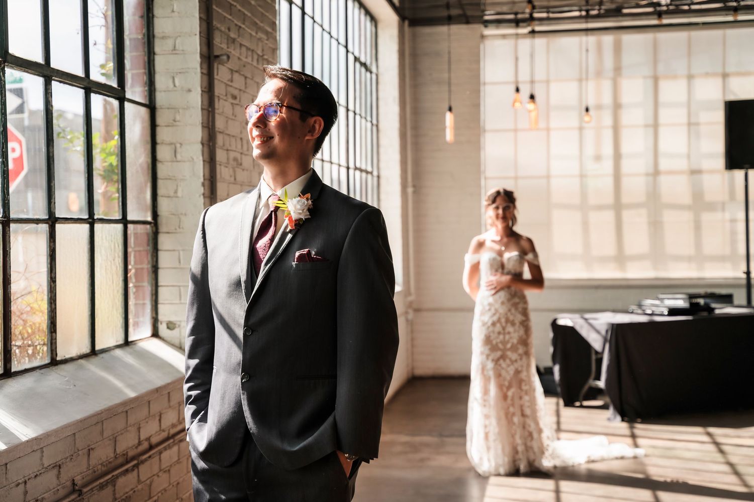 A wedding scene in an industrial venue with exposed brick walls and large windows shows a couple during their first look moment.