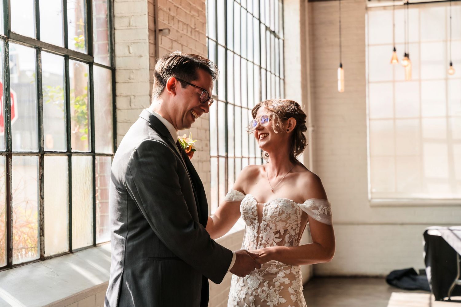 A couple shares a tender moment near large industrial windows in a bright, airy venue.