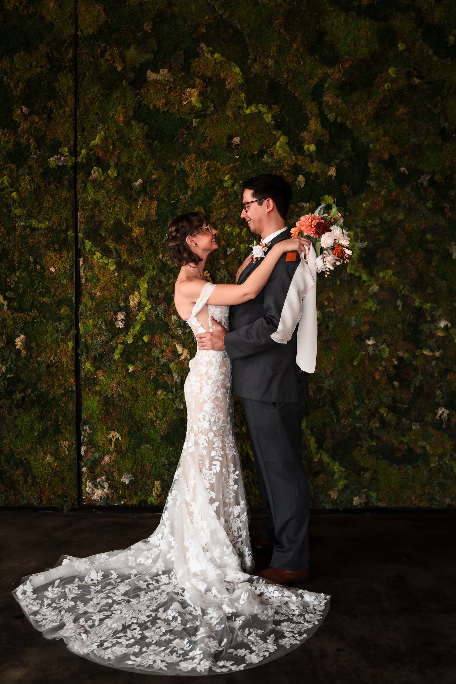 A romantic wedding couple poses against a dark leafy backdrop, with the bride's lace dress trailing elegantly behind.