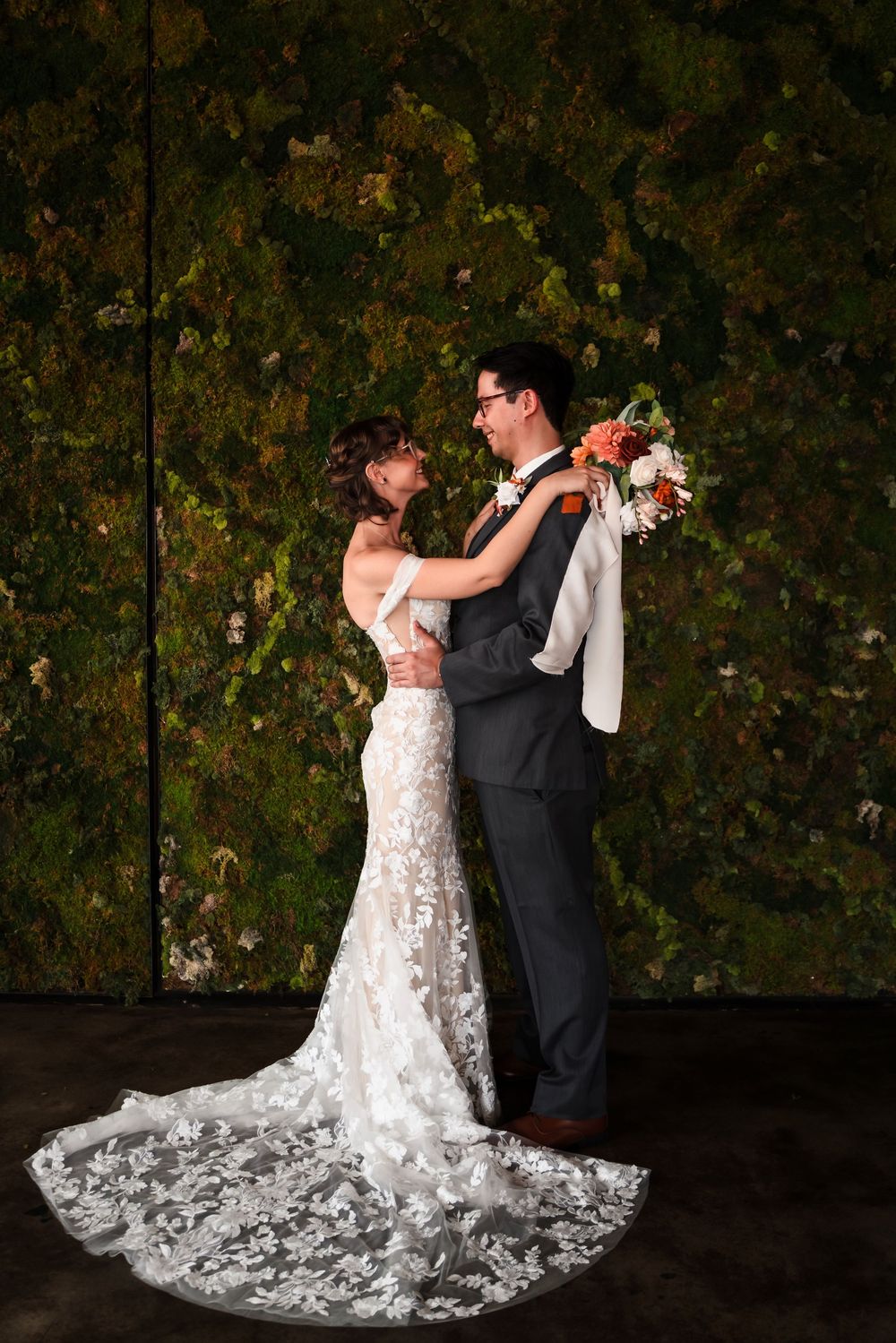 A romantic wedding couple poses against a dark leafy backdrop, with the bride's lace dress trailing elegantly behind.