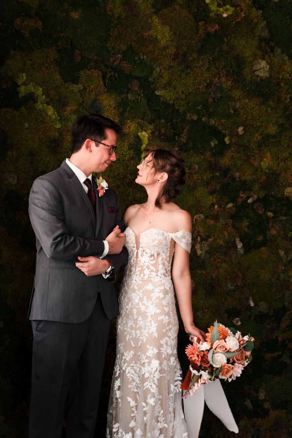 A couple in wedding attire share a romantic moment against a dark green leafy backdrop, with the bride holding a coral bouquet.