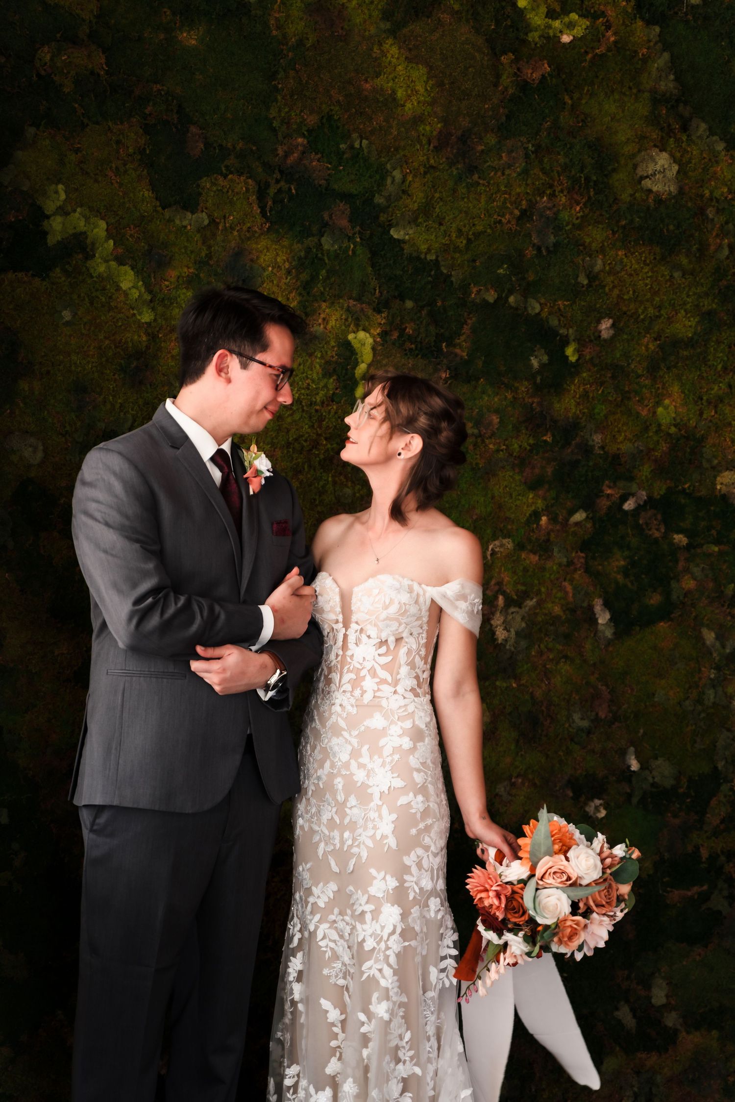 A couple in wedding attire share a romantic moment against a dark green leafy backdrop, with the bride holding a coral bouquet.