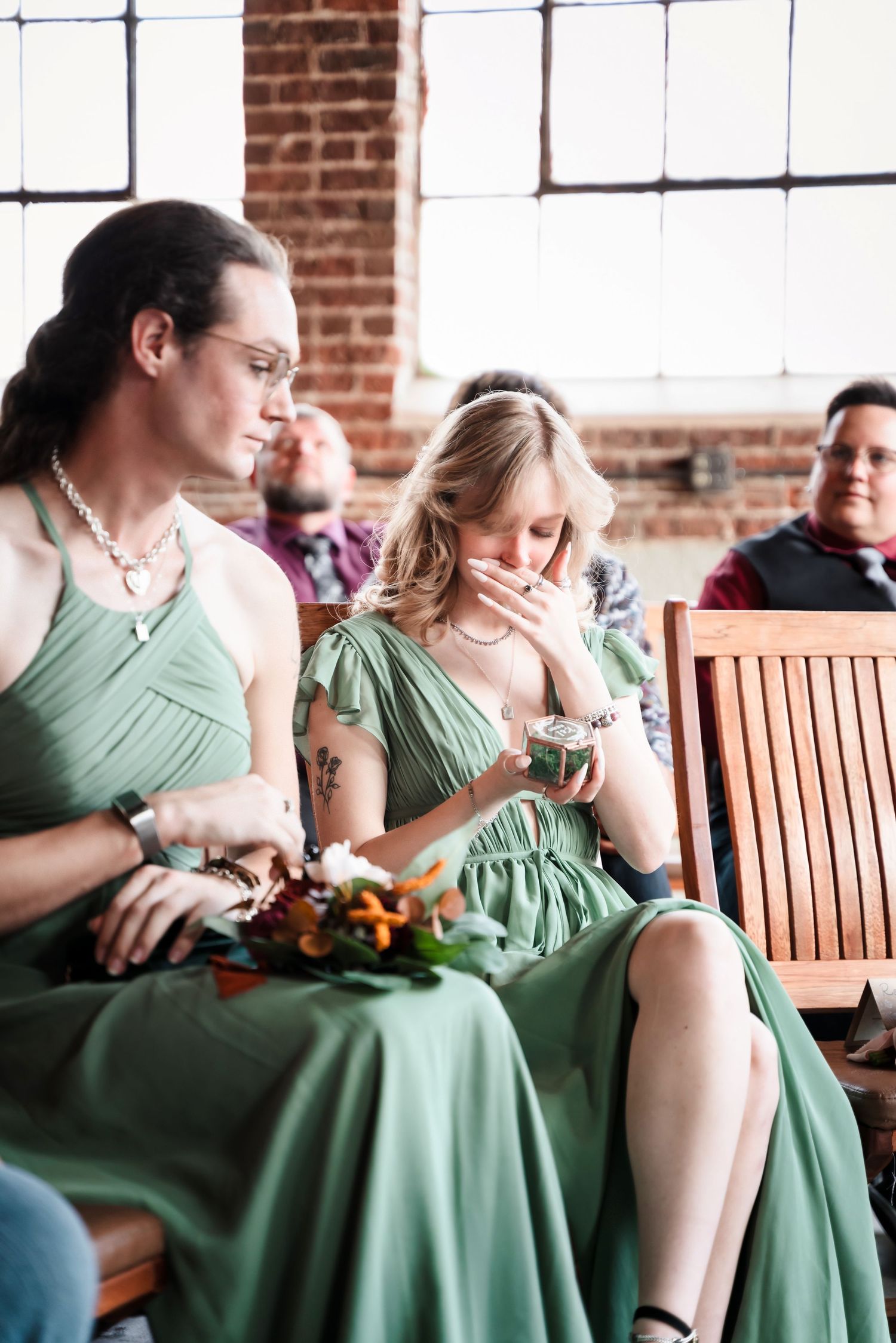 Bridesmaids in sage green dresses sit together on a wooden bench against an exposed brick wall at a wedding venue.