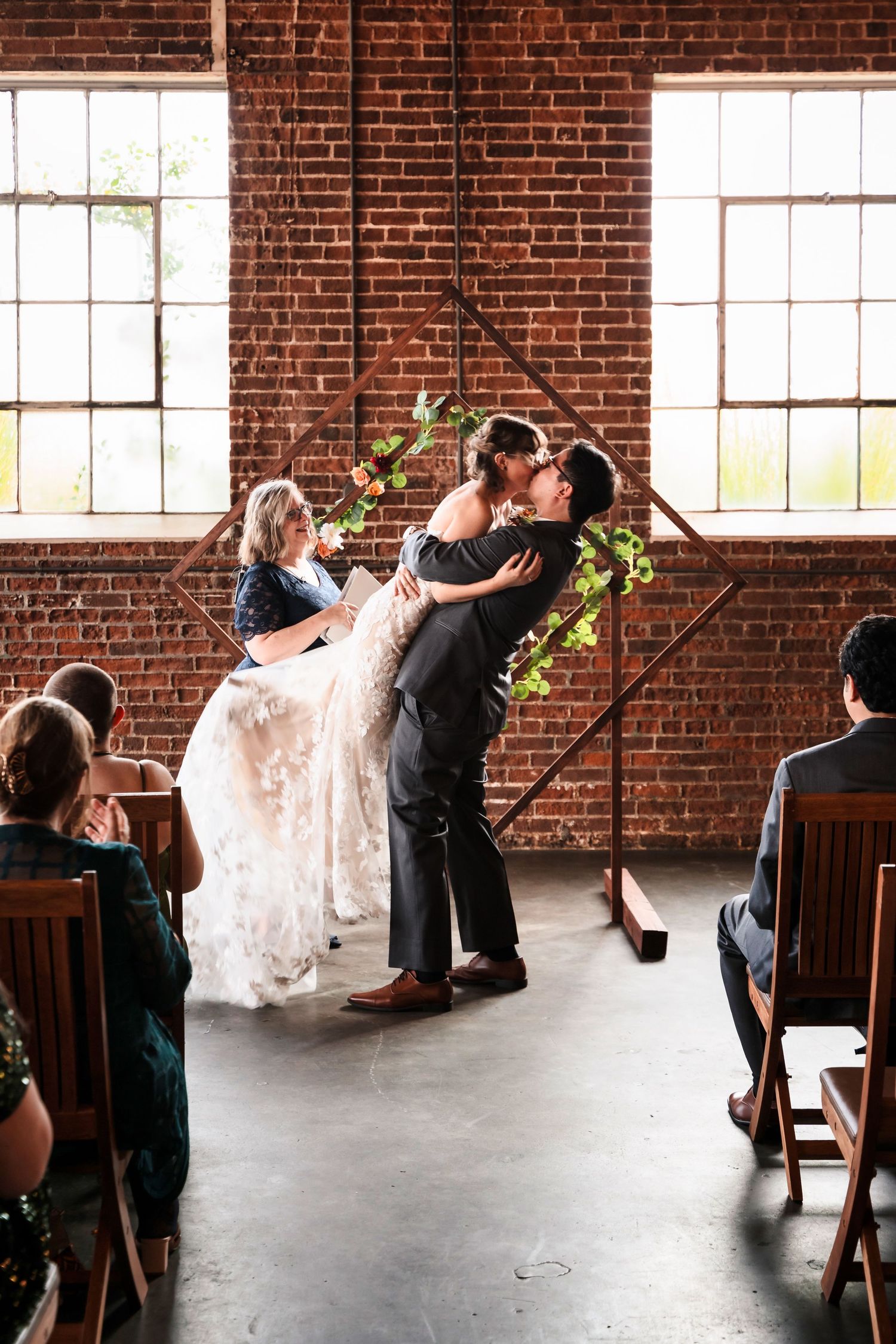 A couple shares their first kiss during a wedding ceremony in an industrial brick venue with large windows and greenery decor.