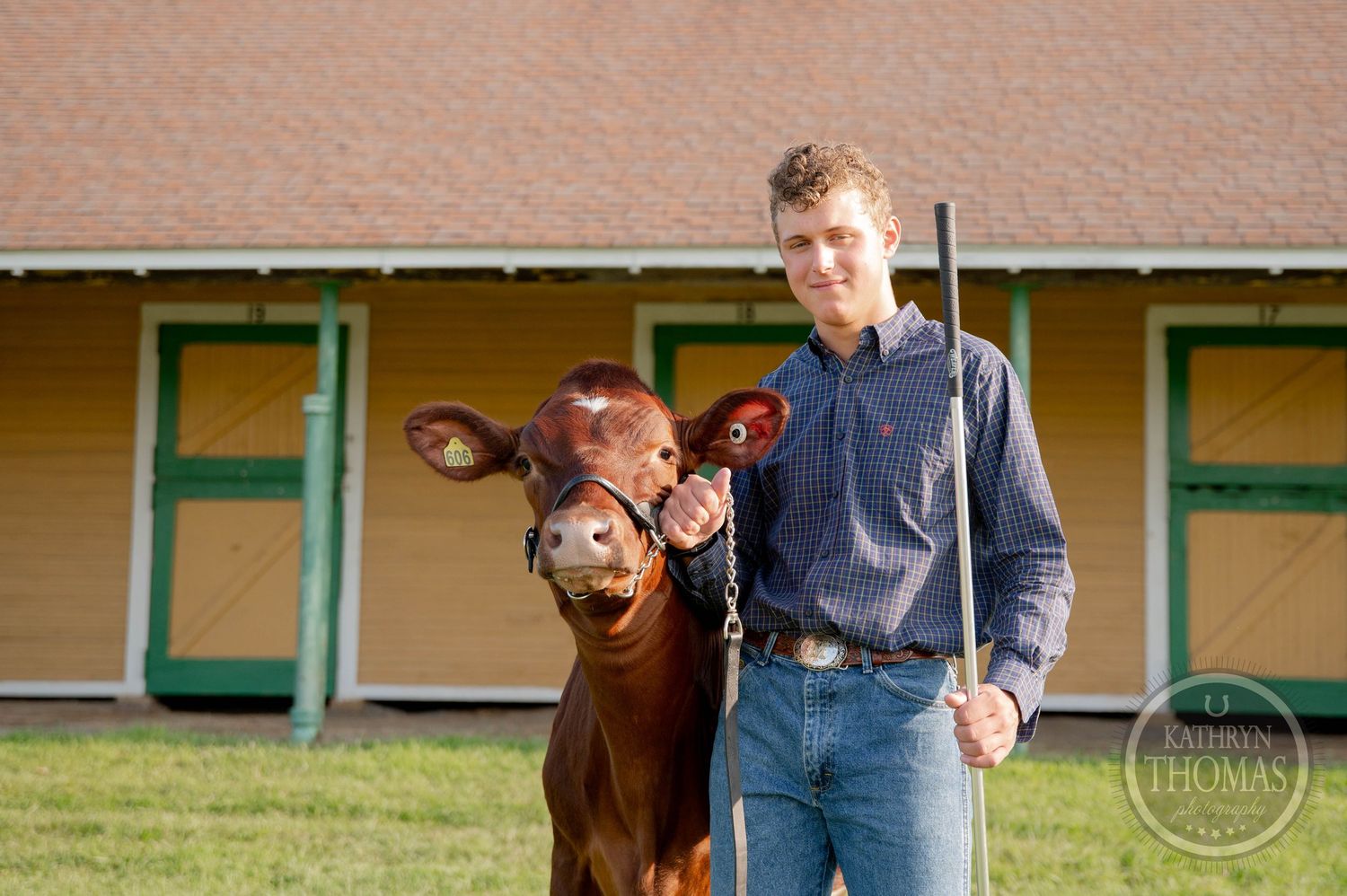 Young man poses with a brown calf at a livestock show, holding a show stick near a yellow barn.