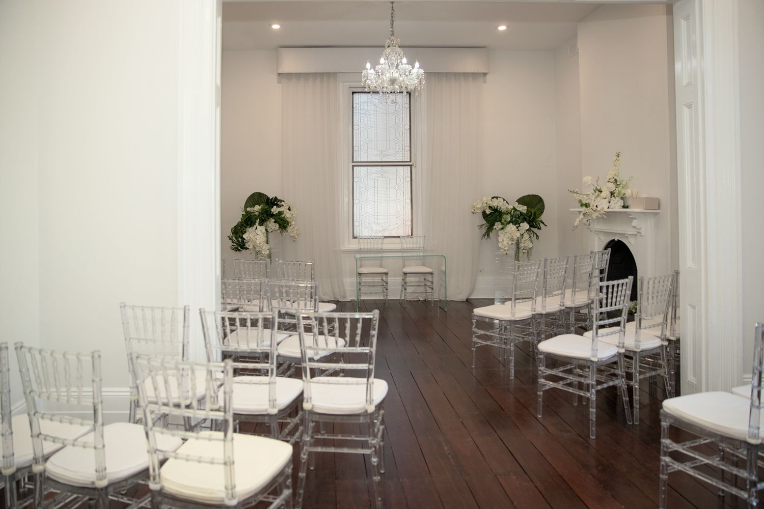 Elegant wedding ceremony room with clear chiavari chairs, white floral arrangements, chandelier, and fireplace on hardwood floors.