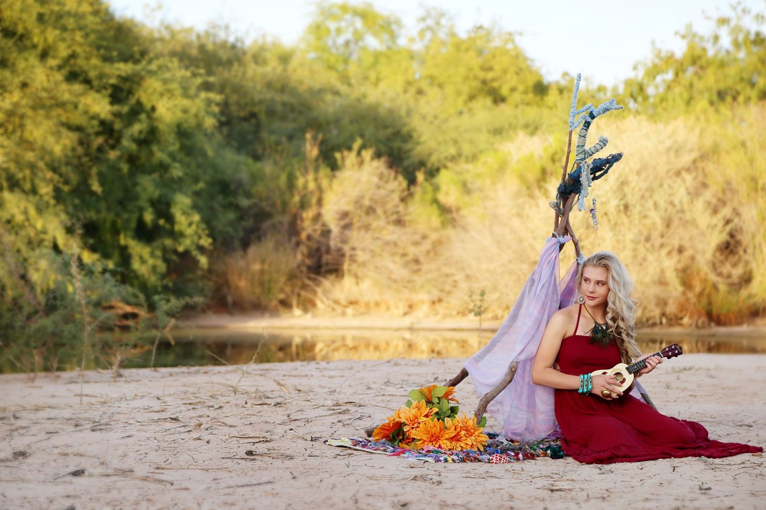 A blonde woman in a red dress plays a small guitar on a sandy riverbank with orange flowers and a wing prop behind her.